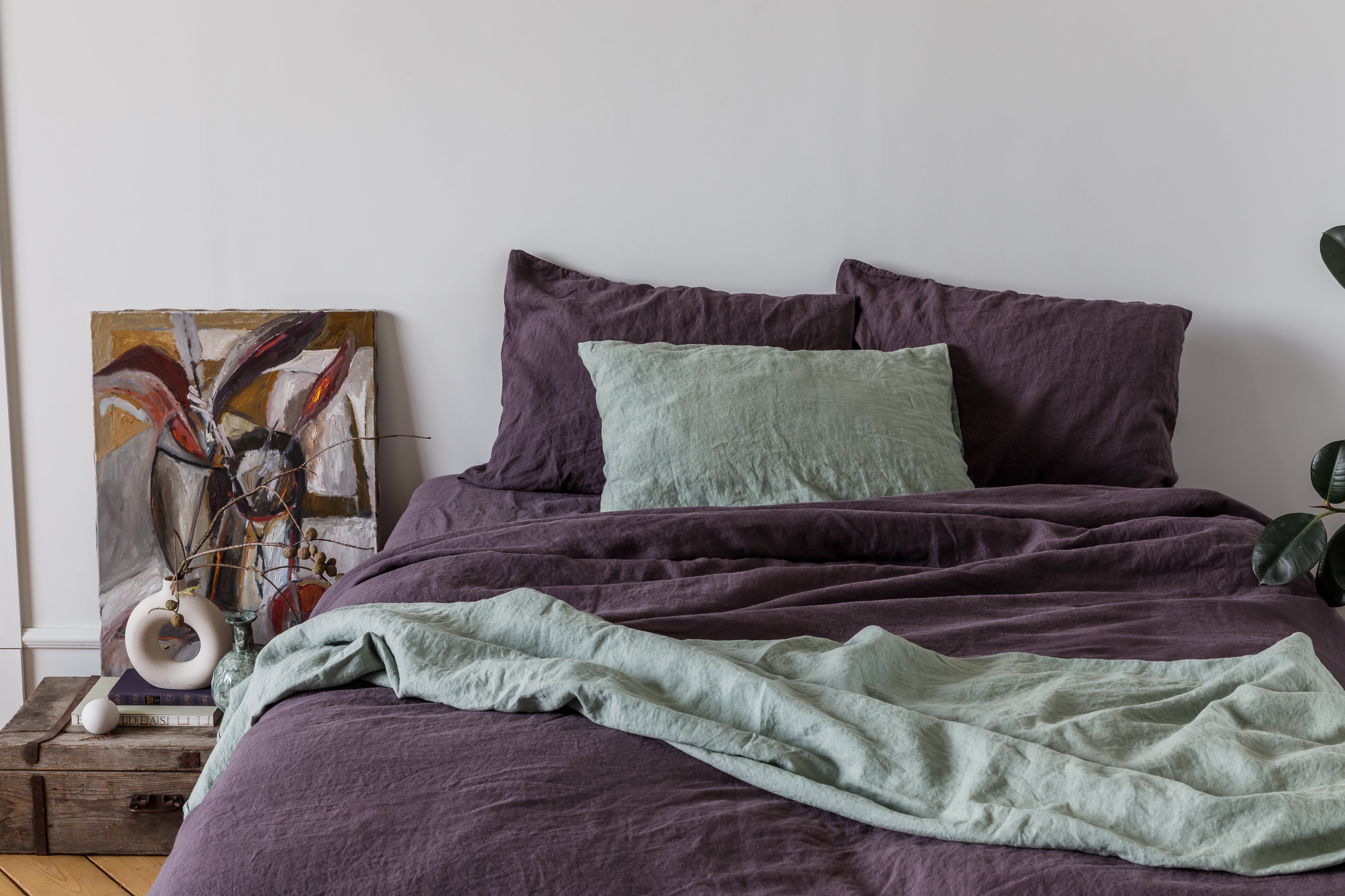 Bed with stonewashed purple linen bedding and green throw blanket in a room with a painting on the wall.