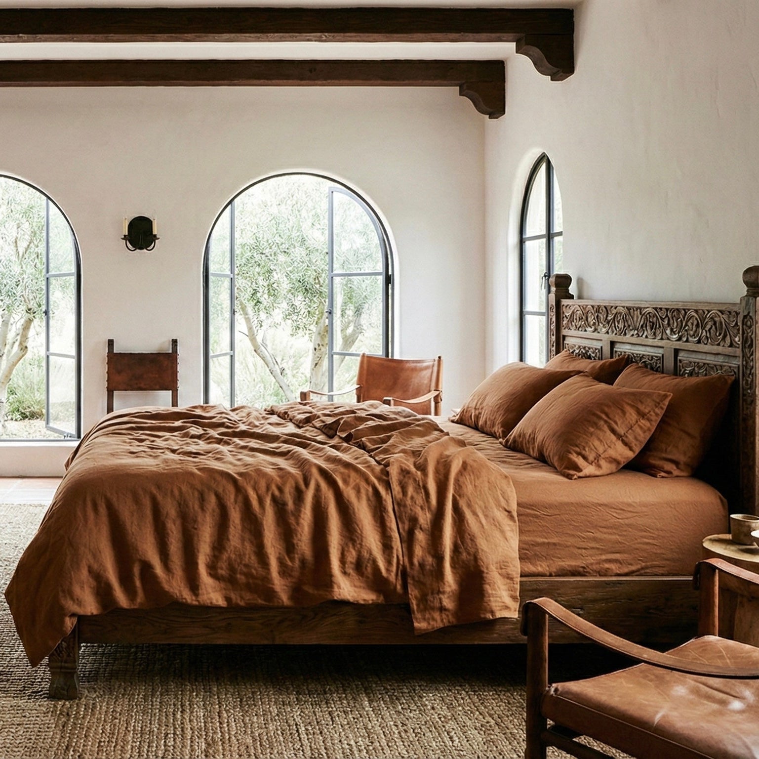 Cozy Mediterranean Californian style bedroom with a bed, fireplace, and large windows. The bed has a linen bedding set of duvet cover and 4 pillowcases in cinnamon brown color