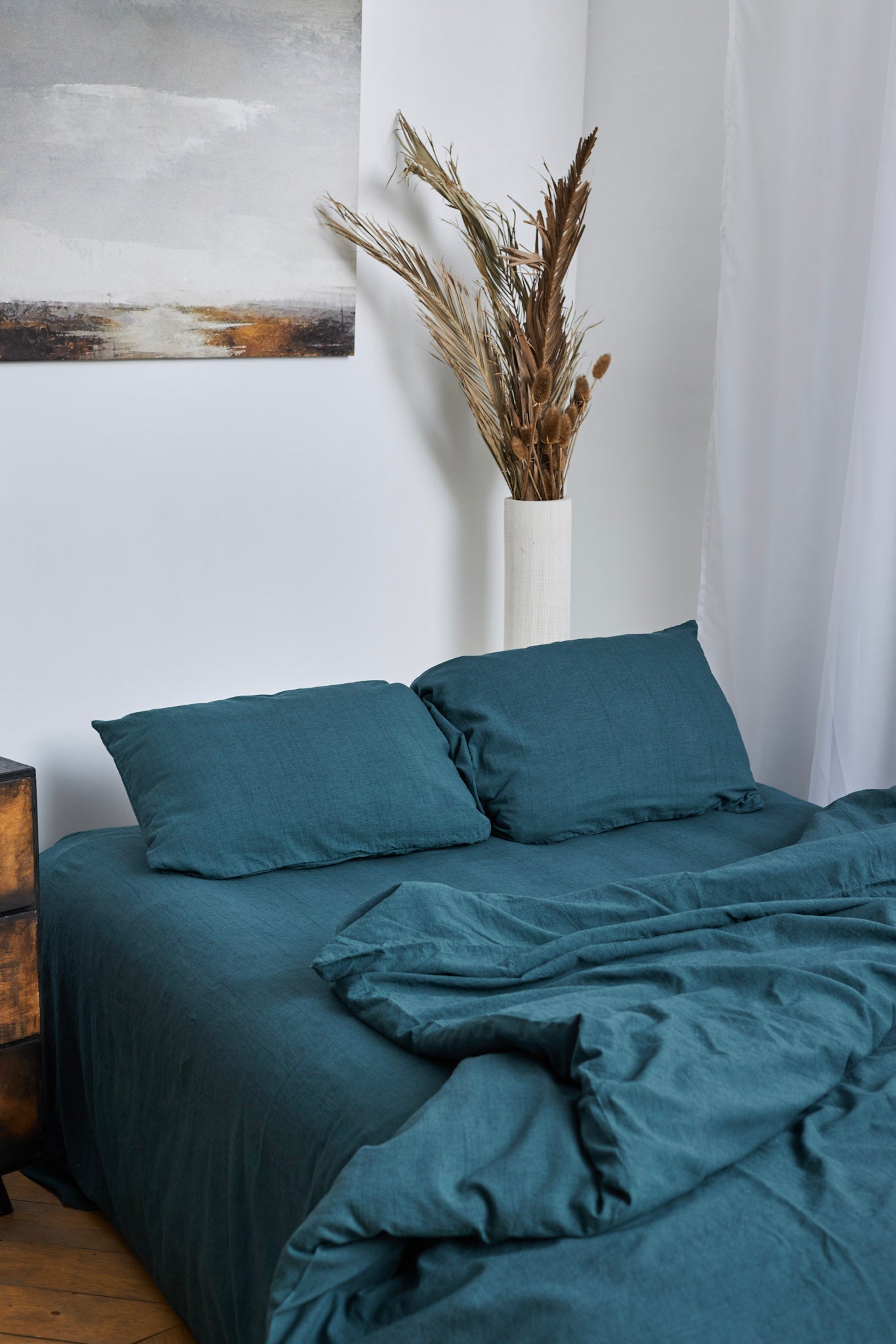 Teal linen bedding set with pillows on a bed with a plant and painting in the background.