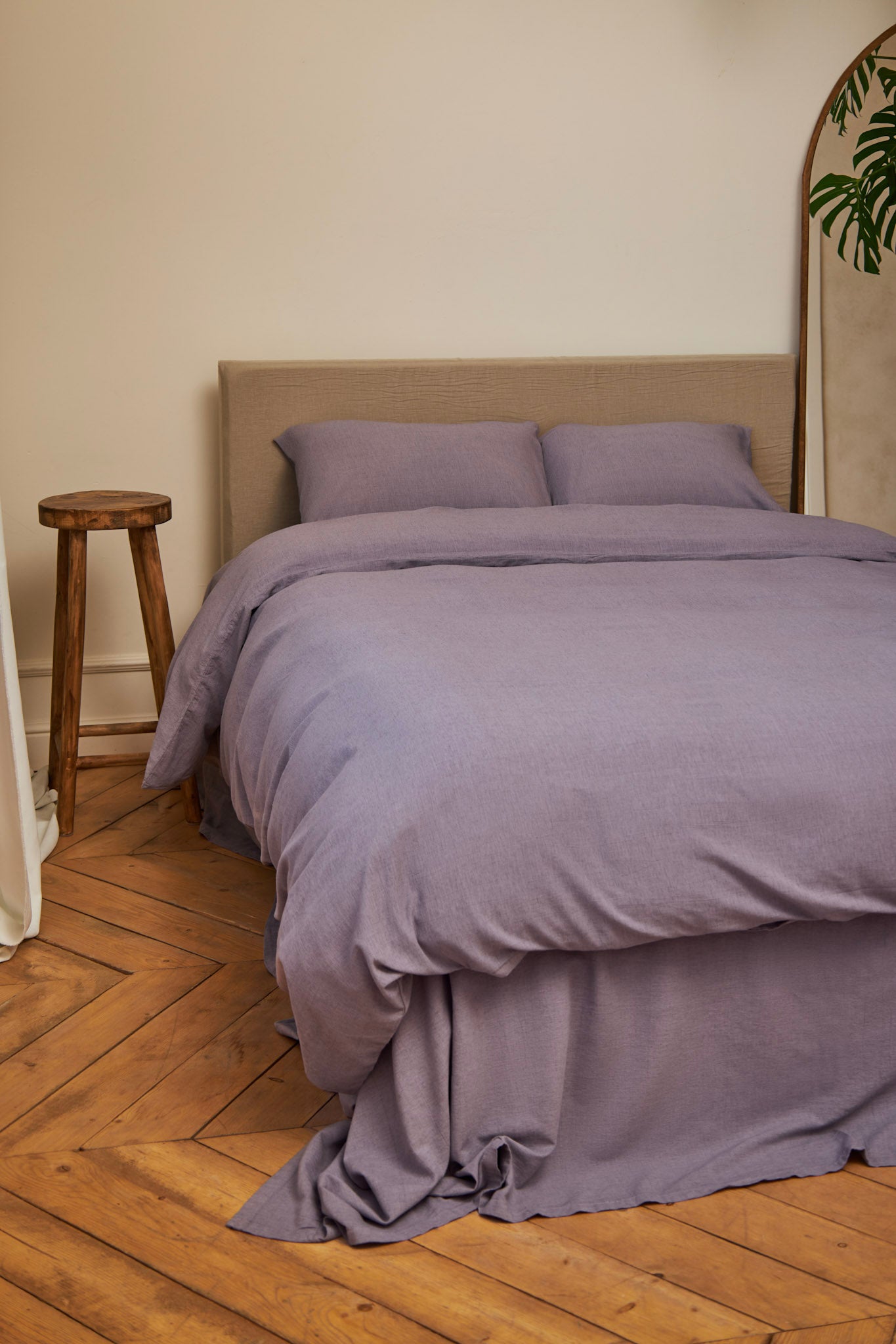 Bed with lavender purple linen bedding set on a wooden floor in a French style bedroom with a mirror and stool.