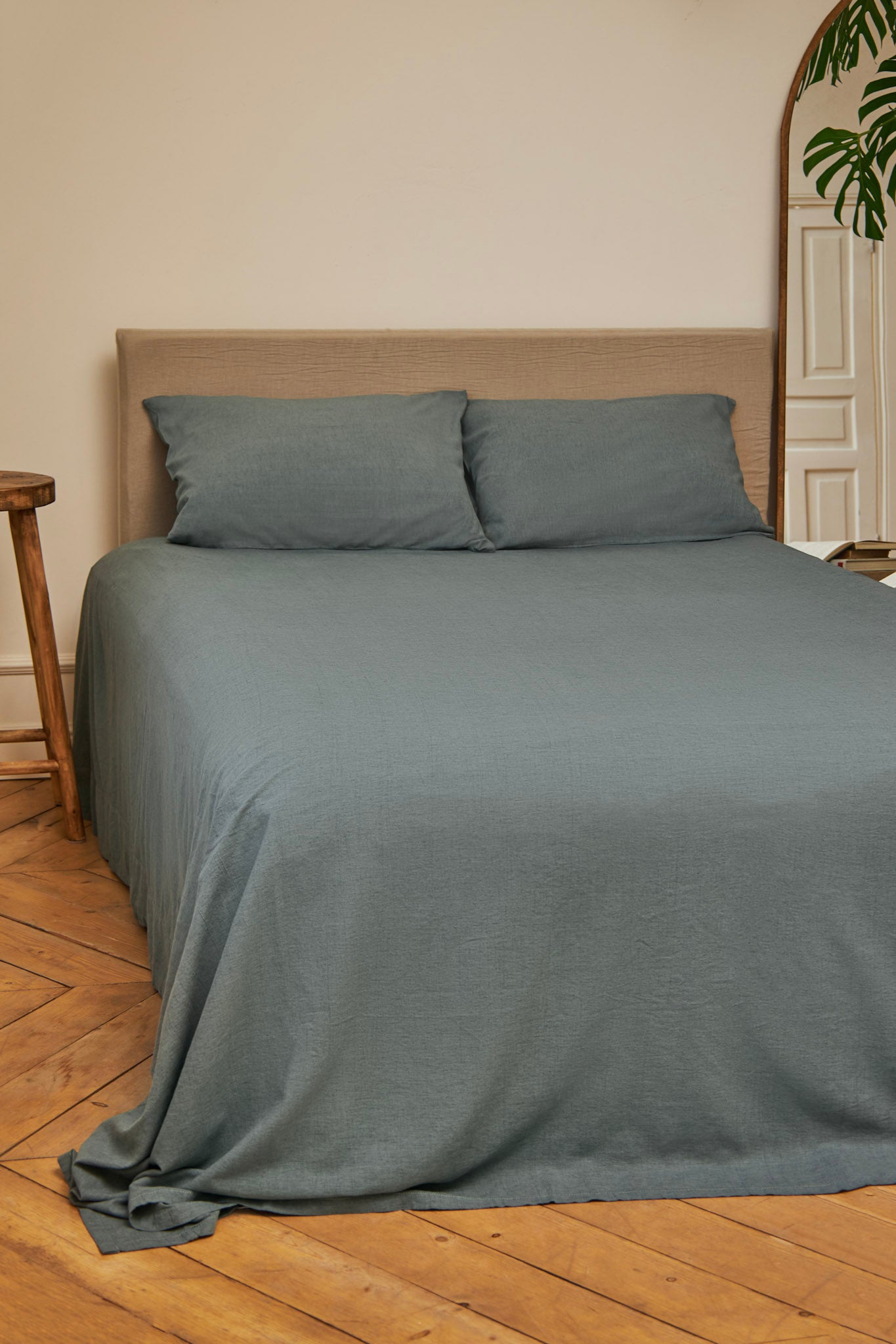 Gray blue linen flax bedspread and pillows on a bed in a room with wooden flooring and a plant.