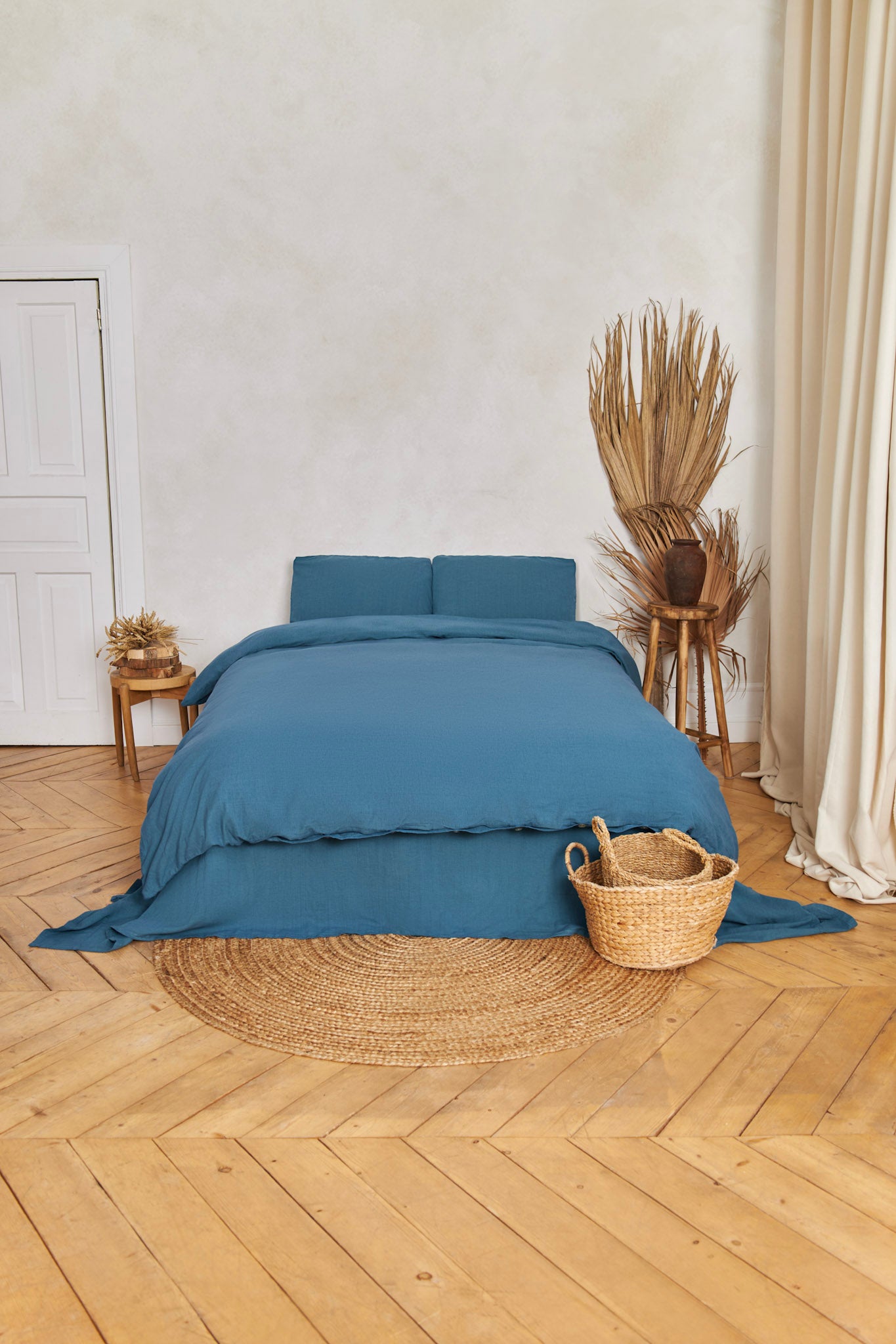 Blue stonewashed linen bedding set on a bed in a room with wooden flooring and decorative elements.