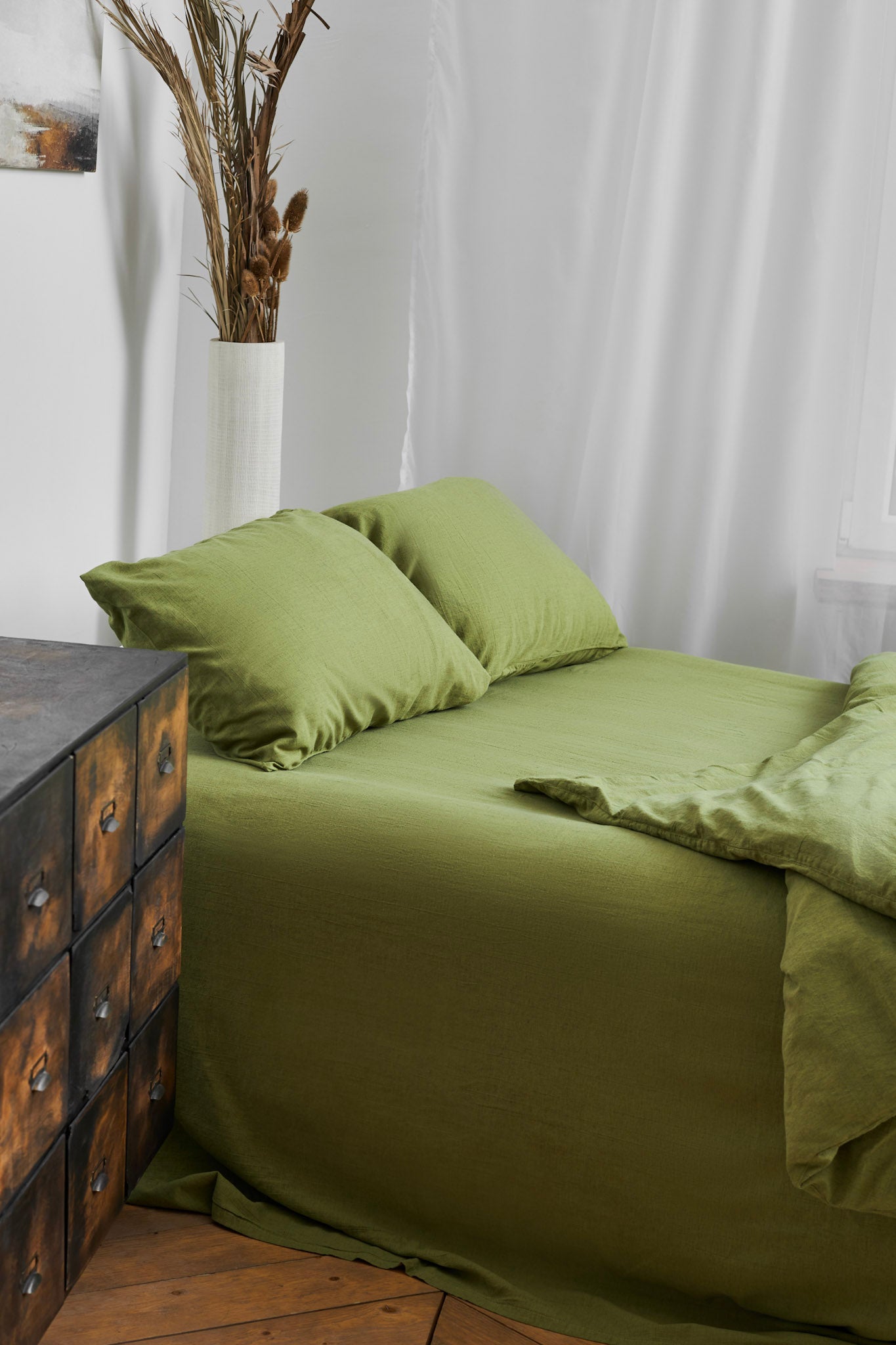 Green linen bedding set on a bed in a room with white curtains and a wooden dresser; modern classic design